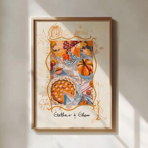 May include: Framed art print depicting a still life with a pie, pumpkins, and autumn leaves. The artwork uses warm colours, including orange, yellow, and blue. The text "Gather & Glow" is at the bottom of the print.