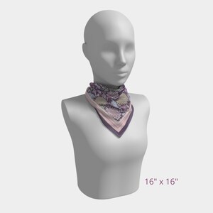 May include: A square scarf, 16 x 16 inches, featuring a floral pattern in shades of purple, pink, and cream. The scarf is displayed on a mannequin and has a contrasting border in darker purple and pink. The overall design is elegant and feminine.