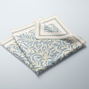 May include: A folded white cotton napkin with a blue and gold floral pattern. The napkin has a border with a gold and blue stripe.