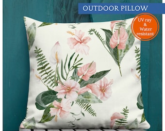 Pode incluir: Almofada quadrada para exterior com um padrão floral de flores de hibisco rosa, antúrios brancos e folhas verdes sobre um fundo creme. A almofada está rotulada como "OUTDOOR PILLOW" e é resistente aos raios UV e à água.
