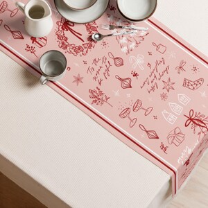 Op de afbeelding: Een roze kersttafelloper met een witte rand en rode accenten. De loper heeft feestelijke illustraties van ornamenten, cadeaus en de tekst "To the reason to be joyful, Merry everything and happy always."