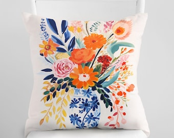 May include: Square decorative pillow with a vibrant floral design. The pillow features a bouquet of orange, pink, and yellow flowers in a blue and white vase. The background is white, and the pillow is placed on a white chair.