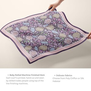 May include: A square scarf with a floral pattern in shades of purple, pink, and blue. The scarf features a border with a pink stripe and is held up by hands. The design includes flowers and fan shapes.
