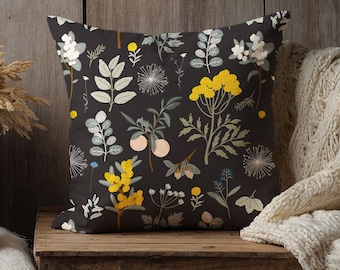 May include: Square throw pillow with a dark gray background and a botanical pattern. The design features yellow, white, and blue flowers, along with green leaves and peach-colored fruit. The pillow is placed on a wooden surface.