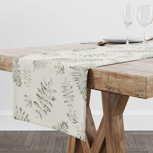 Puede incluir: Camino de mesa color crema con un patrón repetido de hojas de helecho verdes. El camino de mesa está colocado sobre una mesa de madera rústica, con la vajilla visible en el fondo. El diseño añade un toque de naturaleza a la mesa.