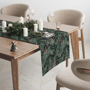Op de afbeelding: Een donkergroene tafelloper met een herhalend patroon van dennennaalden, dennenappels en rode bessen. De loper ligt op een houten tafel, versierd met kaarsen, groen en een bordenset. De scène roept een feestelijke sfeer op.
