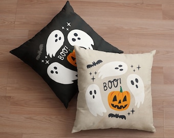 Può includere: Due cuscini quadrati a tema Halloween. Uno è nero con fantasmi bianchi, una zucca e la scritta "BOO!". L'altro è beige con grafica simile. Entrambi hanno accenti di pipistrelli e stelle.