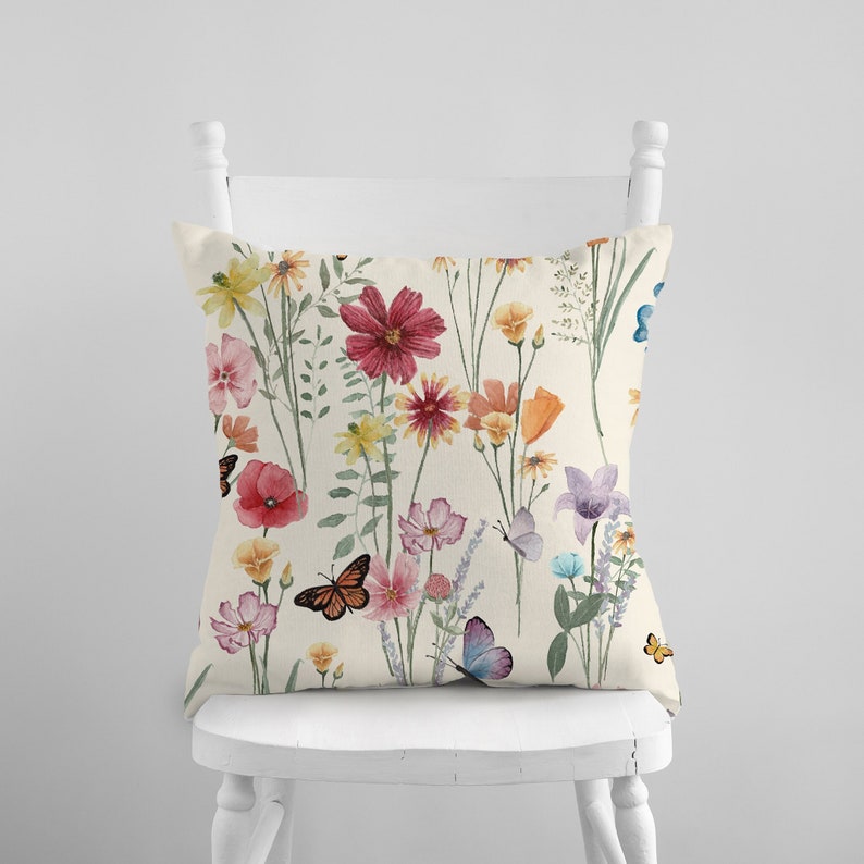 May include: A cream-colored throw pillow with a floral pattern featuring pink, red, orange, and yellow flowers and butterflies. The pillow is sitting on a white chair.