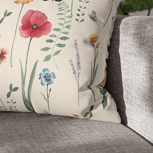 May include: A white throw pillow with a floral pattern featuring red poppies, blue flowers, yellow flowers, and green leaves. The pillow is on a gray couch.