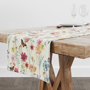 Può includere: Un runner da tavolo bianco con un motivo floreale con farfalle e vari fiori in tonalità di rosa, giallo, blu e arancio. Il runner è posto su un tavolo di legno con due bicchieri da vino e un piatto.