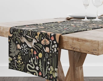May include: A black table runner with a whimsical forest design featuring a running hare, mushrooms, snails, and other flora and fauna. The runner is draped over a wooden table, adding a touch of nature-inspired decor to the dining space.