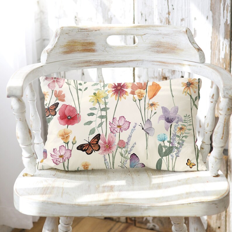 May include: A white pillow with a floral and butterfly print. The pillow features a variety of flowers in shades of pink, red, orange, yellow, and purple, as well as butterflies in shades of blue and orange. The pillow is sitting on a white wooden chair.