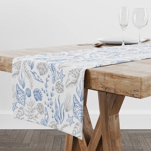 Może przedstawiać: Biały bieżnik stołowy z niebieskim i szarym wzorem muszli i wodorostów. Bieżnik leży na drewnianym stole z dwoma kieliszkami do wina i talerzem.