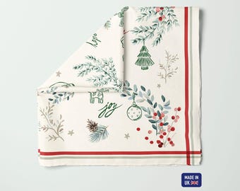 Könnte beinhalten: Cremefarbene Weihnachtstischdecke mit festlichem Design. Mit Illustrationen von Tannenzweigen, Ornamenten und Beeren in Grün, Rot und Grau. Die Worte "hope" und "joy" sind auf dem Stoff geschrieben.