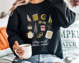 May include: A black crewneck sweatshirt featuring a graphic print of a feather, book, moon, glasses, herbs, candles, scissors, teapot, moth, and a card with the text Stay wild Moon child. The sweatshirt is worn with blue jeans.