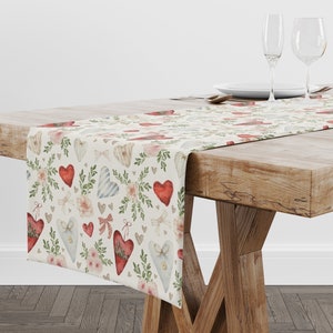 May include: A white table runner with a repeating pattern of red, blue, and white hearts, flowers, and leaves. The runner is on a wooden table with two wine glasses and a plate.