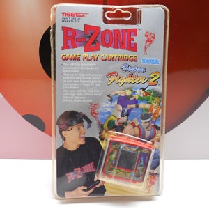 May include: A red and black handheld video game console with a screen showing a fighting game. The console is packaged in a clear plastic clamshell with a white background. The packaging features the text "R-Zone" and "Sega Virtua Fighter 2".