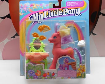May include: A pink My Little Pony toy with a rainbow mane and tail. The pony is holding a small pink brush and is standing in front of a vanity set with a mirror and a flower-shaped seat. The packaging is pink and yellow with the words "My Little Pony" and "Sun Sparkle" on it.