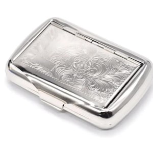 May include: A silver cigarette case with a hinged lid, featuring an engraved floral pattern. The rectangular case has rounded edges and a clasp closure. The design is set against a white background. The case is likely made of metal.