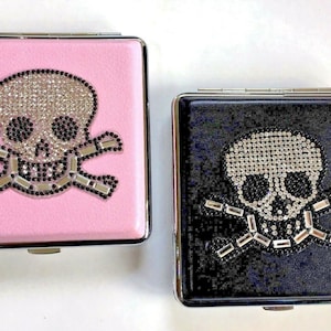 May include: Two compact mirrors featuring a skull and crossbones design. The skull is adorned with rhinestones, while the crossbones are black. One mirror has a pink background, and the other has a black background.