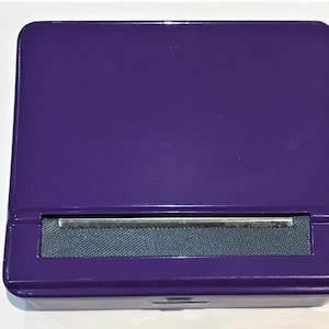 May include: A rectangular purple rolling machine featuring a smooth, glossy finish. The device includes a dark grey fabric rolling apron positioned between two metal bars, designed for manual operation to create uniform cylindrical items.