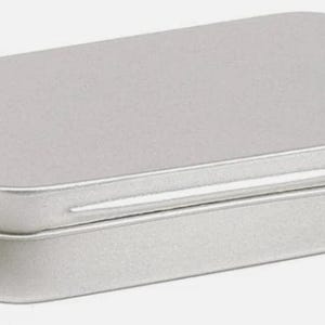 May include: A small, rectangular silver tin with a rounded lid. The tin is made of metal and has a simple, minimalist design. The lid fits snugly onto the base, creating a secure closure. This container is suitable for storing small items.