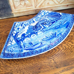 May include: A blue and white porcelain dish with a unique, curved shape. The dish features a detailed landscape scene with bridges, boats, and figures. The rim is decorated with a patterned border. The overall design is a classic, vintage style.