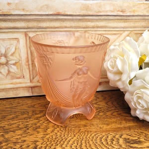 May include: A peach-coloured, Art Deco-style glass vase with a detailed relief of a woman. The vase has a wide, rounded bowl and a pedestal base. The design includes floral and draped elements. The vase is displayed on a wooden surface.