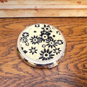 May include: A circular silver compact mirror featuring a floral pattern. The flowers are black and white, set against a pale yellow backdrop. The compact is open, revealing a mirror. It rests on a wooden surface.