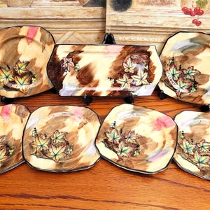 May include: A collection of decorative ceramic plates and a rectangular serving dish. The plates and dish have a brown, beige, and pink watercolour design with green and yellow leaf accents. The edges are outlined in black.