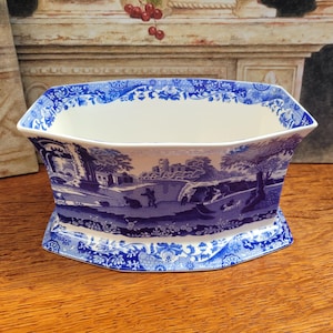 May include: A blue and white ceramic planter with a landscape design. The planter has a rectangular shape with a flared rim and a decorative base. The landscape scene depicts trees, figures, and a building.