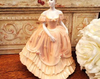 May include: A porcelain figurine of a woman in a peach-colored ball gown with ruffled layers and floral accents. The figurine has a delicate face and is posed gracefully. The background features a decorative wall and a bouquet of white flowers.