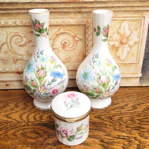 Op de afbeelding: Driedelige porseleinen set met twee vazen en een kleine pot. Het witte porselein is versierd met kleurrijke bloemmotieven, waaronder roze rozen, blauwe korenbloemen en andere wilde bloemen. De vazen en de pot hebben een gouden rand.