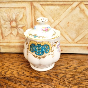 Puede incluir: Un azucarero de porcelana blanca con tapa, con detalles florales y dorados. El azucarero tiene un diseño turquesa y dorado en la parte delantera. La tapa está rematada con un pequeño pomo decorativo. El azucarero mide unos 10 cm de alto.