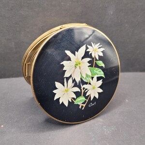 May include: A round, vintage compact with a black enamel top. The top features a floral design of white poinsettias with green leaves. The compact has a gold-tone metal frame and a hinged opening. The artist's signature, "Carl", is visible.