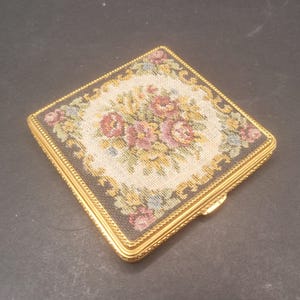 Op de afbeelding: Een vierkant compact met een gouden rand en een bloemenmotief in tapisserie. De tapisserie heeft een centraal motief van roze en paarse bloemen omringd door groene bladeren op een crèmekleurige achtergrond.