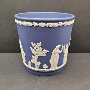 May include: A blue ceramic planter with white relief detailing. The planter features a band of leaves around the rim and classical figures and trees around the body. The planter is cylindrical and has a slightly flared top.
