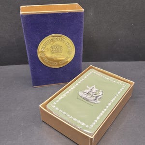 May include: A deck of playing cards in a tan box with a blue velvet case. The card box has a gold emblem that reads "WADDINGTONS FINE PLAYING CARDS". The cards have a green background with a white floral border and a silver illustration.
