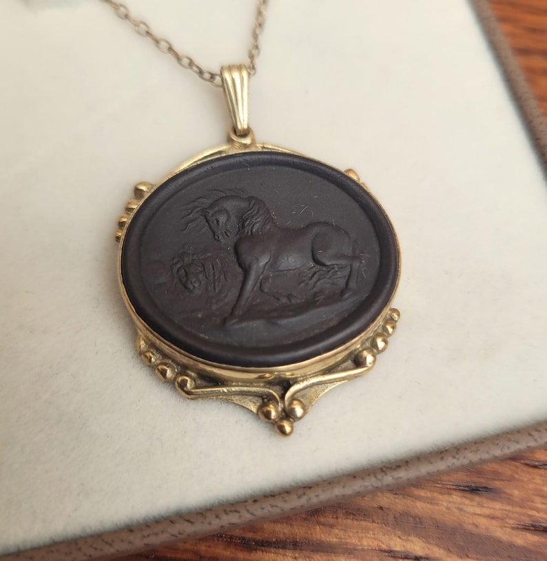 May include: Gold-toned pendant necklace featuring a dark oval cameo with a detailed relief of a horse. The pendant is framed in gold and hangs from a delicate chain. The horse is depicted in a classical style.