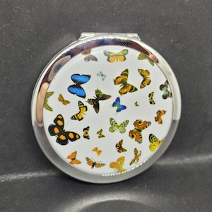 May include: A round, silver compact mirror with a white surface featuring a colorful butterfly pattern. The butterflies are in various sizes and colors, including blue, yellow, orange, and black. The mirror is closed and has a shiny, metallic border.