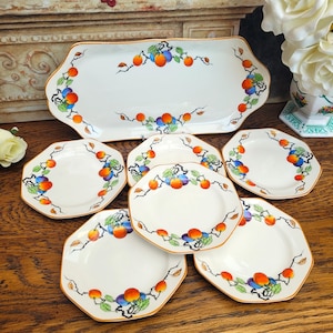 May include: A white ceramic serving platter and six matching plates. Each piece features a decorative design of orange and blue fruit with green leaves and brown stems. The edges of the plates and platter have a gold trim.