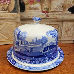 May include: A white and blue ceramic cheese dome with a matching plate. The dome and plate feature a blue pastoral scene with trees, buildings, and figures. The rim of the plate has a blue floral pattern.