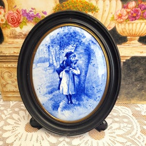 May include: Oval framed porcelain plaque with a blue and white pastoral scene. The artwork depicts two children near a tree. The frame is black with a gold inner rim and a stand on the back.
