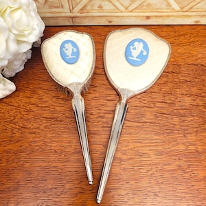 May include: A vintage vanity set featuring a brush and a hand mirror. Both items have cream-colored surfaces with a diamond pattern and a blue cameo design. The handles are silver-toned and have a ribbed design. The set is displayed on a wooden surface.