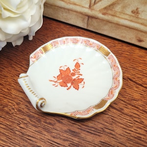 May include: A white porcelain seashell-shaped dish with a floral design in orange and gold accents. The rim features a delicate orange and gold pattern. The dish has a rolled edge and is set on a wooden surface.