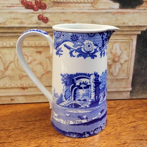 May include: A white ceramic jug with a blue and white pastoral design. The jug features a curved handle and a spout. The blue design includes floral patterns and a landscape scene with a bridge and cows. The jug is a decorative item.