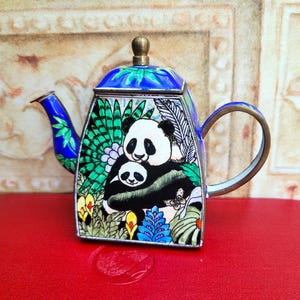 May include: A decorative teapot with a panda bear design. The teapot has a blue lid and spout, a gold handle, and a colorful panda bear illustration on the front. The teapot is sitting on a red surface.