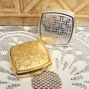 May include: Two square compact mirrors with a geometric pattern. One mirror is gold and the other is silver.