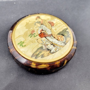 May include: A round, tortoiseshell compact mirror with a gold rim. The mirror has a painted scene of three people in traditional Japanese clothing walking in a landscape.