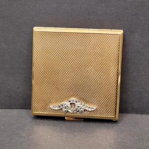 May include: A gold-toned compact with a diamond-shaped design and a textured surface. The compact has a hinged lid and a small mirror inside.